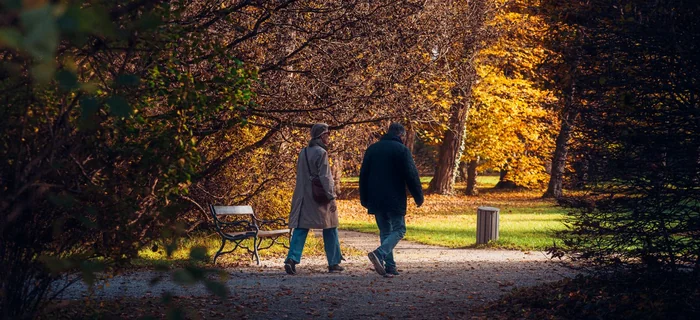Two older adults walking side by side along a leaf-covered path through an autumn park