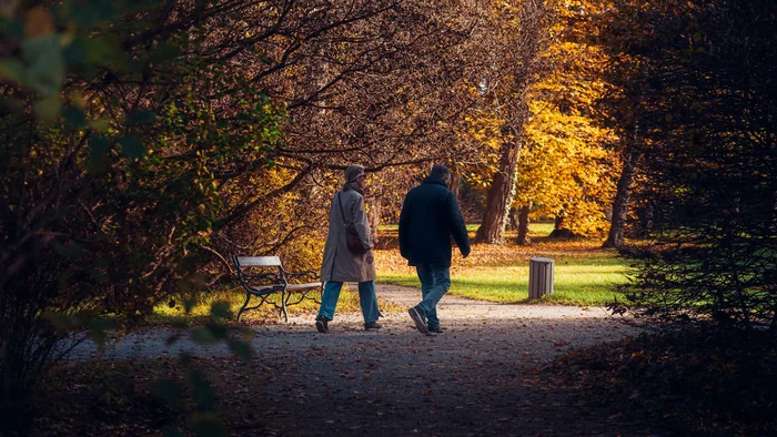 Two older adults walking side by side along a leaf-covered path through an autumn park